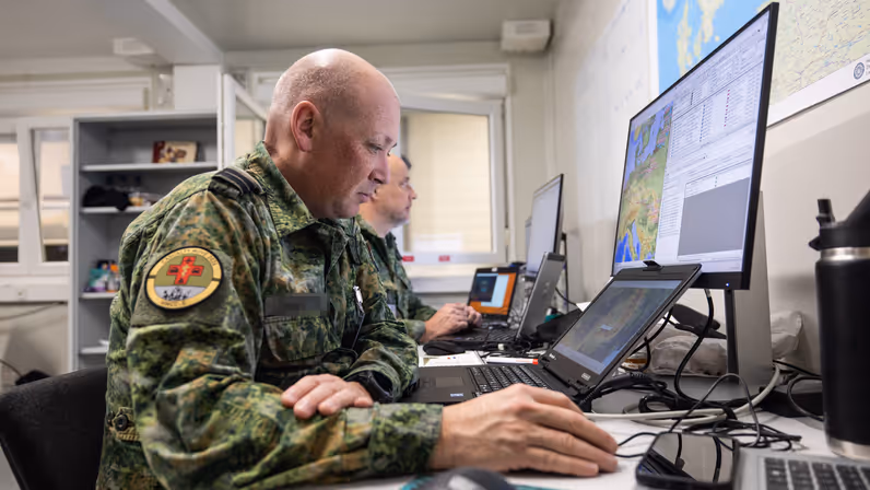 Twee militairen voor meerdere computerschermen aan een bureau. 