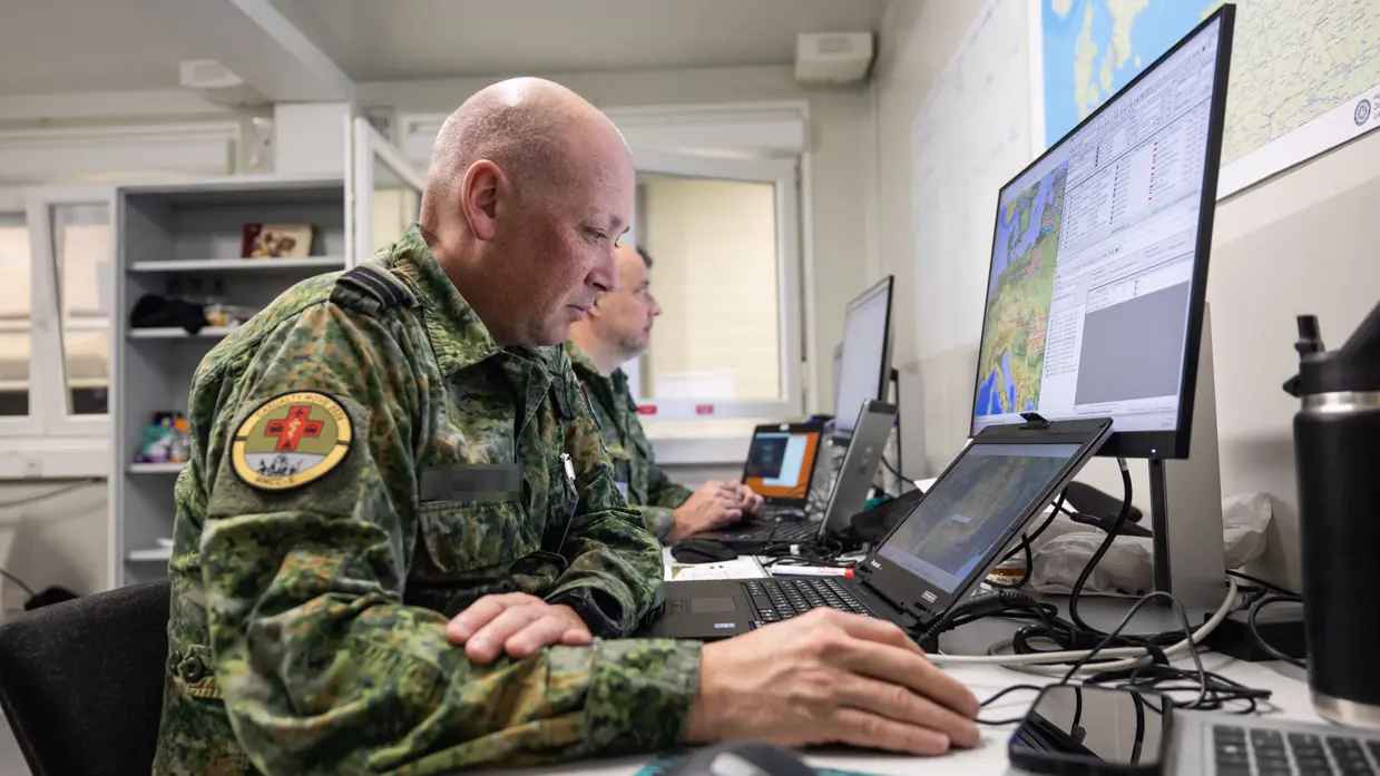 Twee militairen voor meerdere computerschermen aan een bureau. 