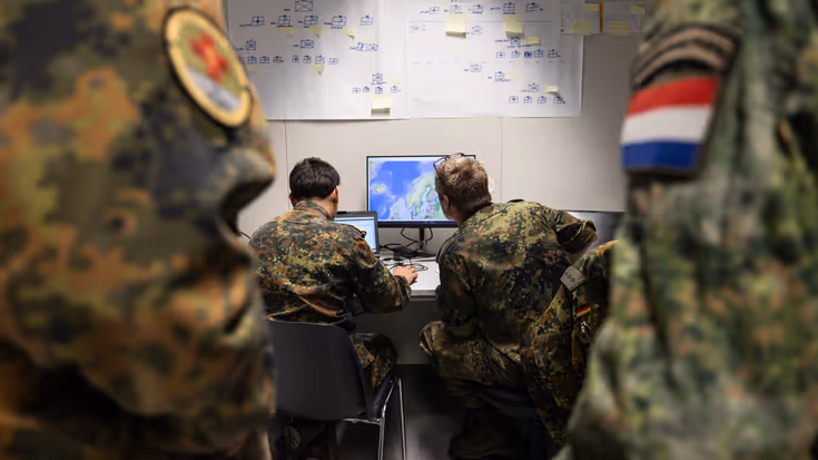 Militairen in uniform zitten achter een computer met kaarten en schema’s op de muur. 