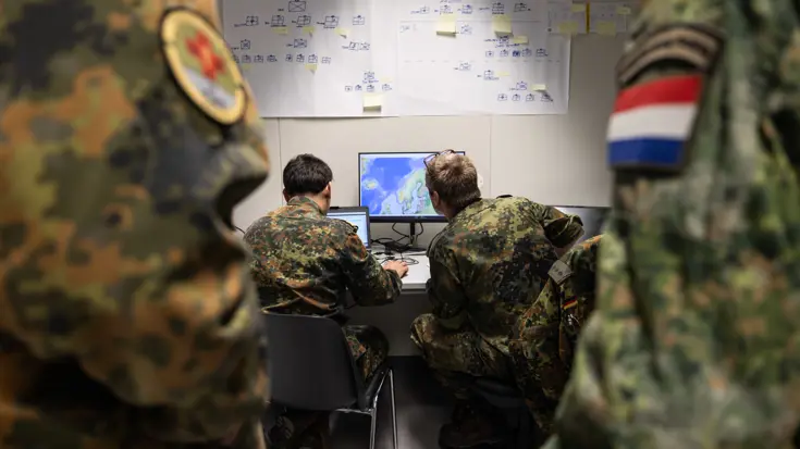 Militairen in uniform zitten achter een computer met kaarten en schema’s op de muur. 