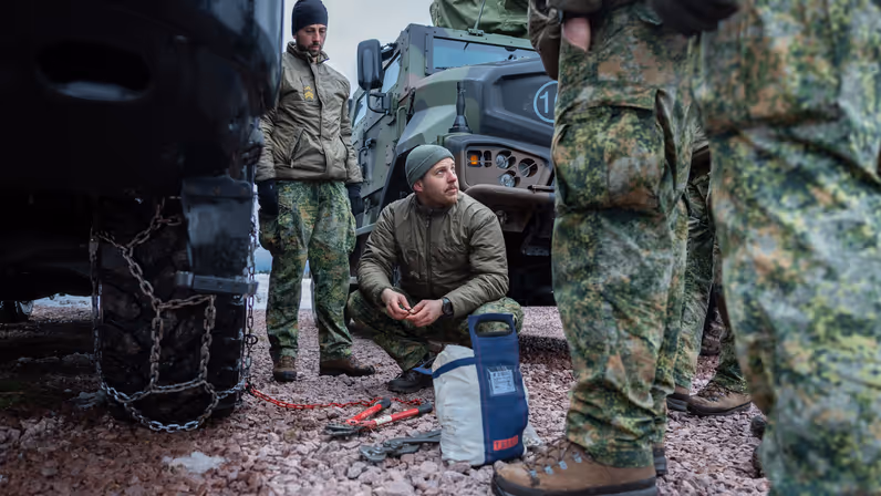 Sergeant Timo geeft instructie over het repareren van een sneeuwketting. In zijn hand een grote tang om schakels dicht te knijpen en om hem heen toekijkende manschappen.