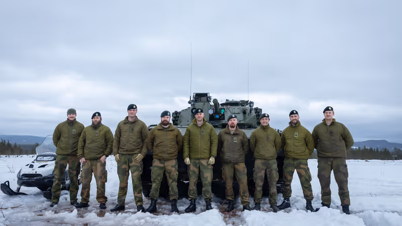 Het Noorse team van instructeurs opgelijnd in de sneeuw, voor een CV90 en een sneeuwscooter. 