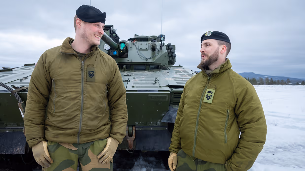 Twee Noorse sergeanten staan voor een CV90 lachend met elkaar te praten.