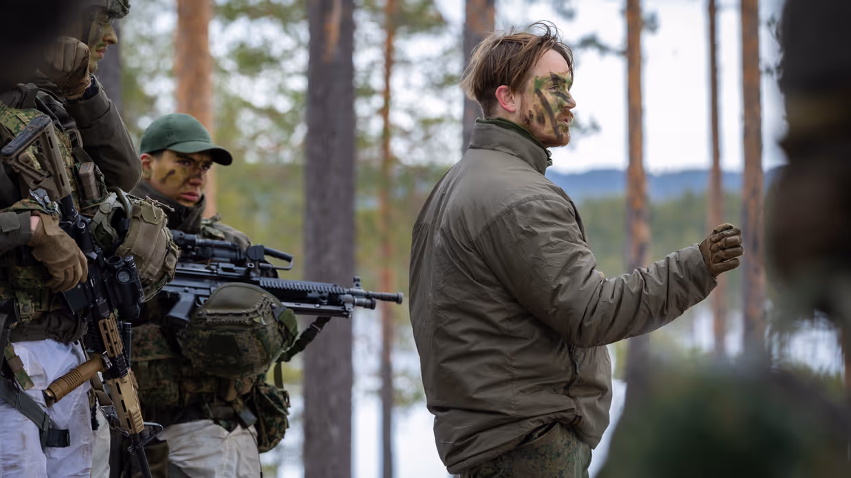 Sergeant Xander geeft in het bos in winterse omstandigheden uitleg aan collega’s. Xander heeft camouflage op zijn gezicht, achter hem staan collega’s in volle wapenrusting.