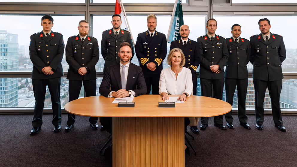 2 mensen zitten aan tafel, achter ze staan reservisten.