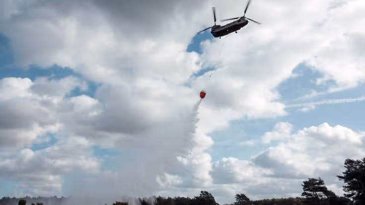 Tegenlichtopname van een Chinook met Bambi Bucket die water loost boven een heidestrook waar met verticale vlaggen een pad is uitgezet.