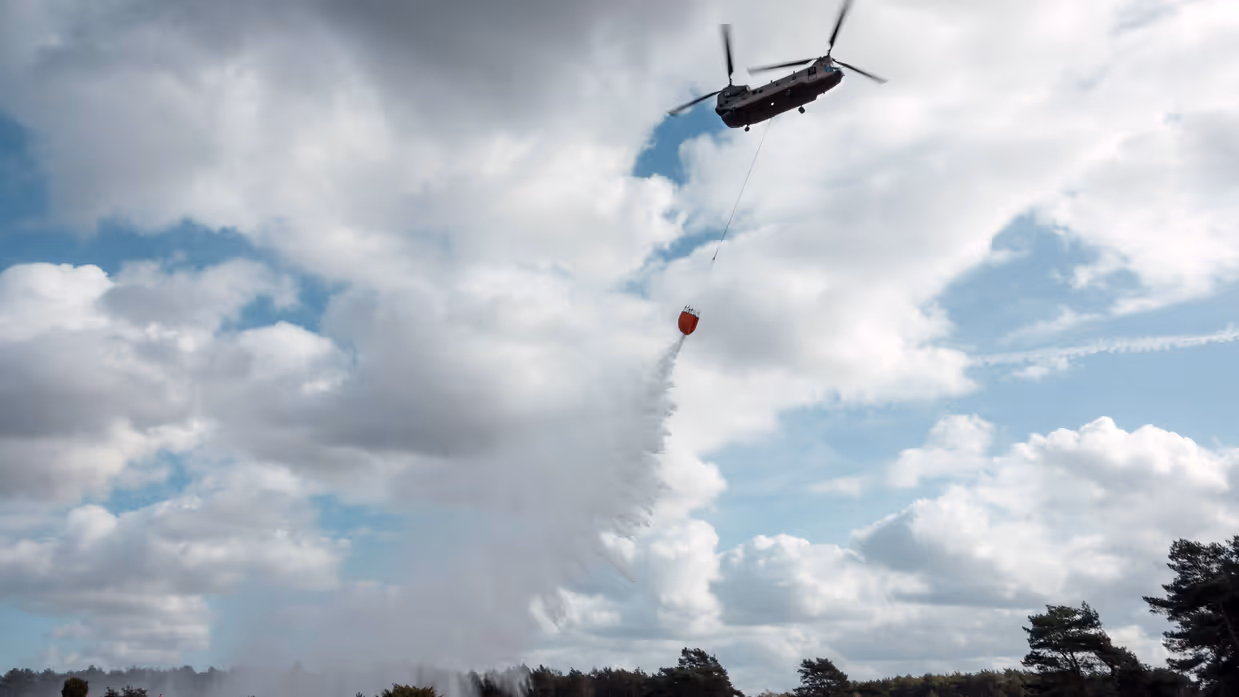 Tegenlichtopname van een Chinook met Bambi Bucket die water loost boven een heidestrook waar met verticale vlaggen een pad is uitgezet.