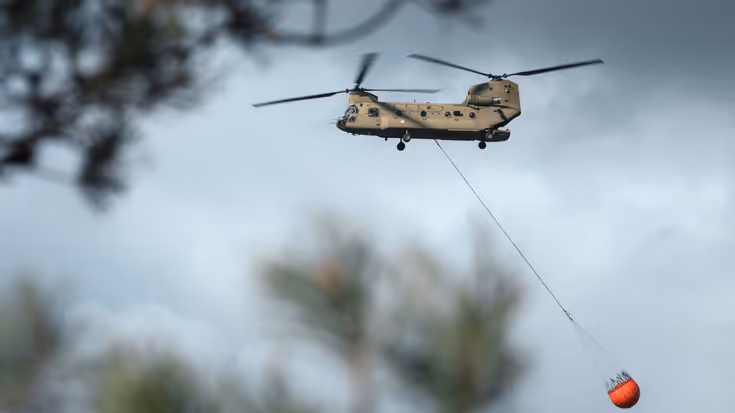 Een Chinook vliegt met een lege Bambi Bucket van rechts naar links. In de voorgrond zijn takken van bomen te zien zodat de helikopter daar tussendoor lijkt te vliegen. Geen bijschrift