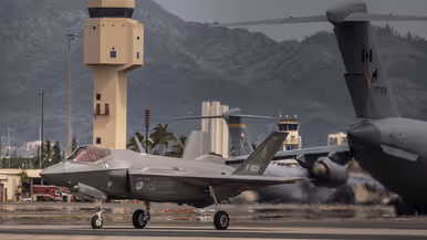 Een F-35 taxiet op Hawaii voor de verkeerstoren langs. In de achtergrond staan C-17 transportvliegtuigen van de VS en uit Canada