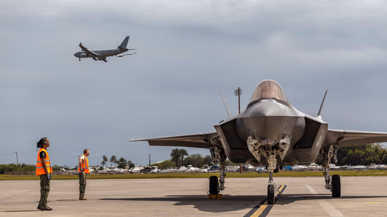 Een F-35 wordt door grondpersoneel met oranje hesjes naar zijn parkeerplaats geleid terwijl daarboven een A330 in de landing is te zien.