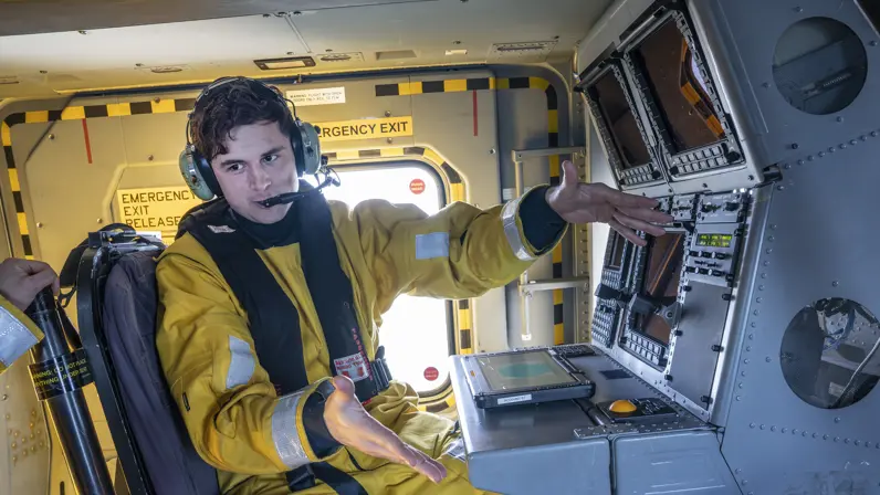LTZ 3 Teun zit in geel dompelpak op de plek van de helicopter sensor operator, met zijn tablet binnen handbereik. Hij legt met weidse armgebaren iets uit.