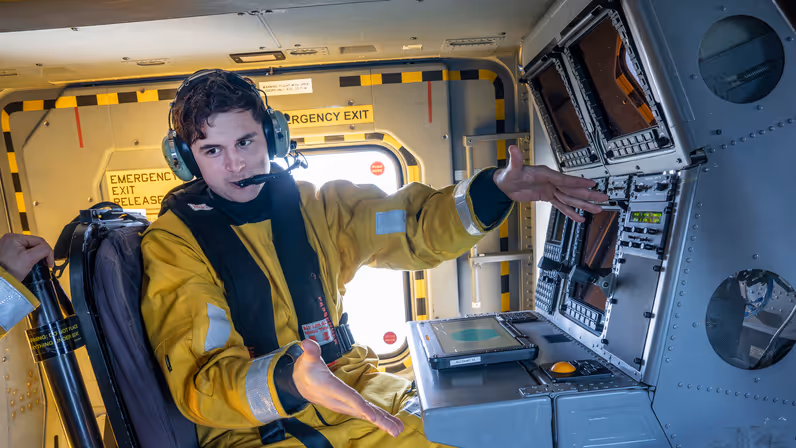 LTZ 3 Teun zit in geel dompelpak op de plek van de helicopter sensor operator, met zijn tablet binnen handbereik. Hij legt met weidse armgebaren iets uit.