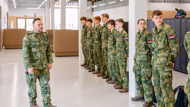 Op een strak witte vloer in een modern licht pand staan jongeren in militaire kleding op een rij. Ervoor loopt sergeant-majoor Tim Nelissen die hen toespreekt.