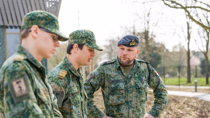 Sergeant-majoor Tim Nelissen in gevechtspak met blauwe baret luistert met zijn handen in zijn zij naar twee jongens in militair gevechtstenue. Ze staan op een verharde weg met groenstroken eromheen.