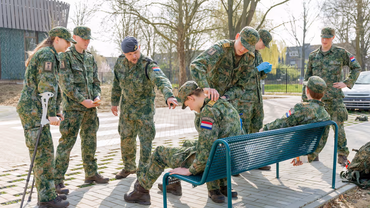 Twee jonge militairen op een bankje met daar omheen vijf andere jonge militairen. Ze dragen allemaal militaire petjes. Een van hen staat op krukken, een ander draagt blauwe latexhandschoenen. Tim praat tegen twee staande militairen en wijst naar een van de militairen op het bankje.