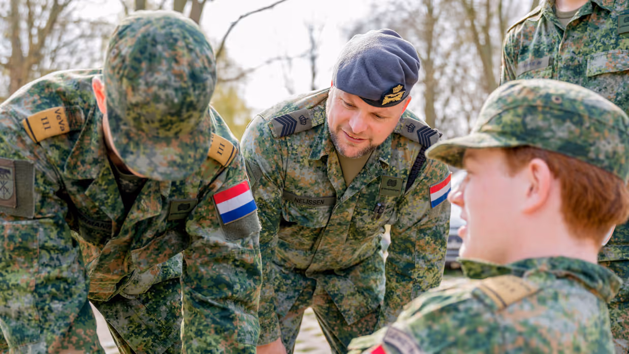 Een jonge militair zittend op een bankje, een andere met blauwe latexhandschoen aan buigt naar hem toe. In het midden van hen kijkt Tim toe.