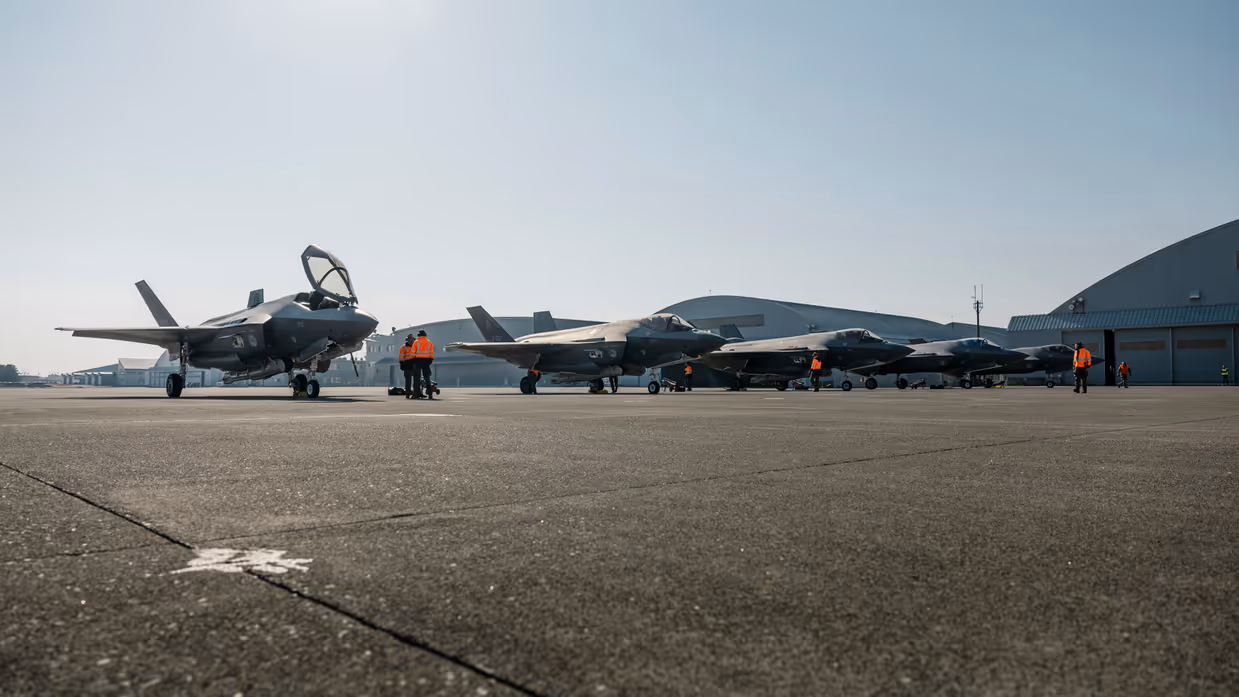 Alt-tekst: 5 F-35’s naast elkaar op een platform met in de achtergrond een aantal ronde hangers. Er staat personeel met oranje jassen tussen de F-35’s. 