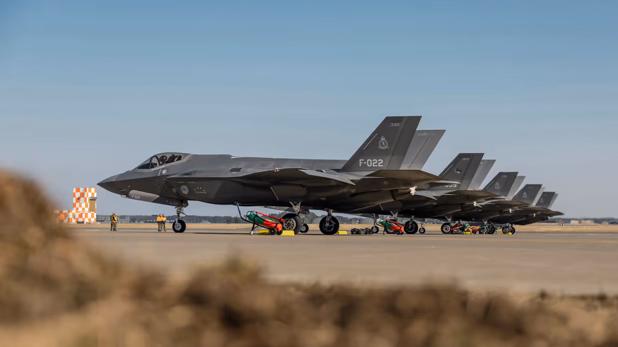 Vijf Nederlandse F-35’s naast elkaar op een platform. Links is een rood-wit geblokt gebouw te zien.