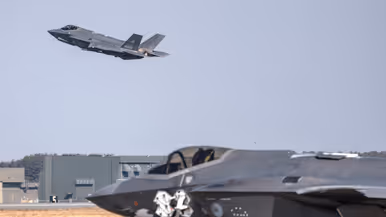 En Japanse F-35 stijgt op met in de voorgrond een Nederlandse F-35 en in de achtergrond een groen gebouw.