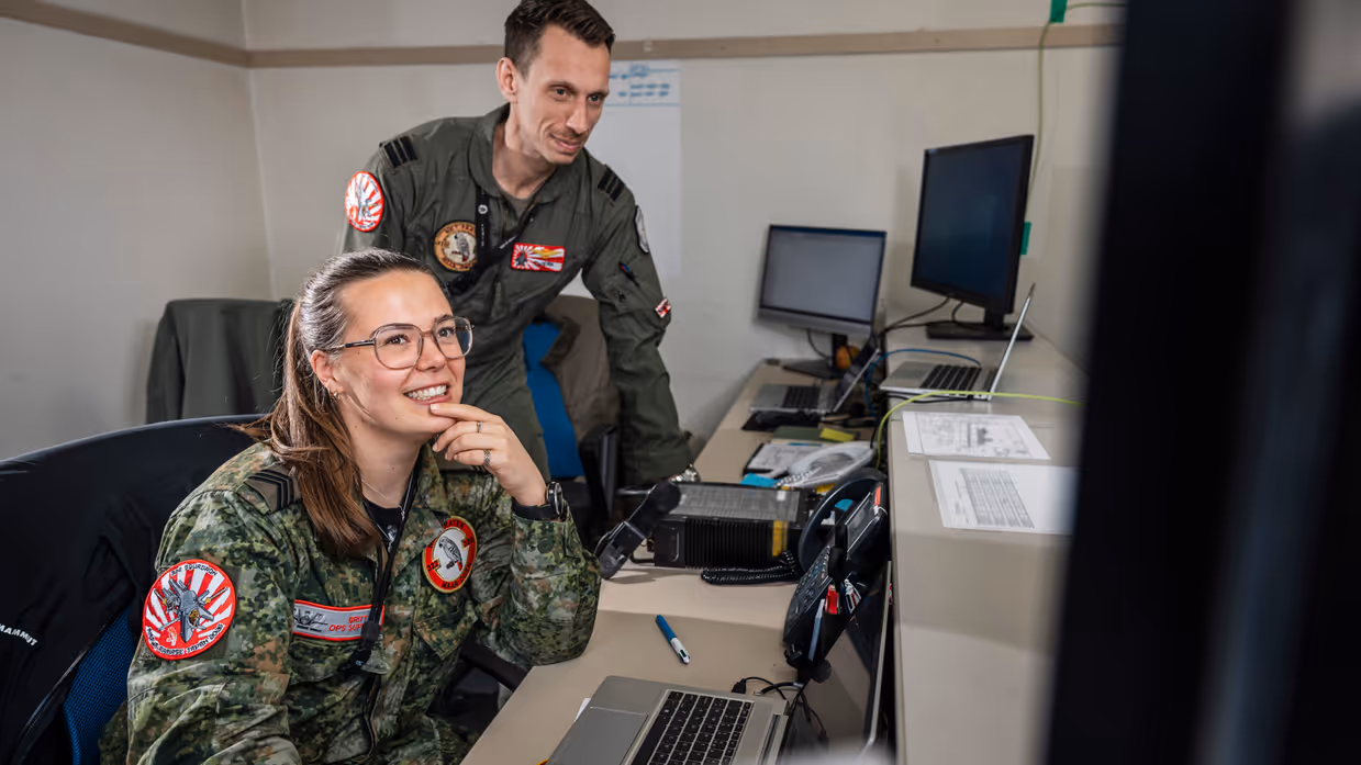 Sergeant Britt zittend in de voorgrond aan een bureau. In de achtergrond een vlieger, staand en over haar heen kijkend.