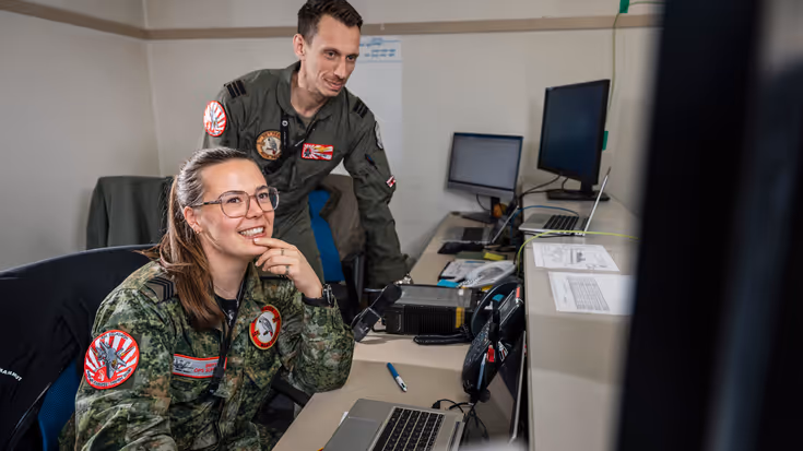 Sergeant Britt zittend in de voorgrond aan een bureau. In de achtergrond een vlieger, staand en over haar heen kijkend.
