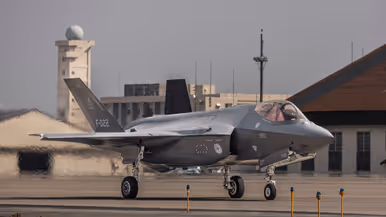 Een F-35 taxiet met in de achtergrond een radartoren, gebouwen en een hangar, en in de voorgrond gele paaltjes met daarop een blauwe lamp.