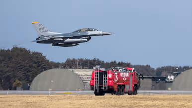 En Amerikaanse F-16 stijgt op met in de voorgrond een brandweerwagen met Japanse opschriften en in de achtergrond ronde vliegtuig-shelters.