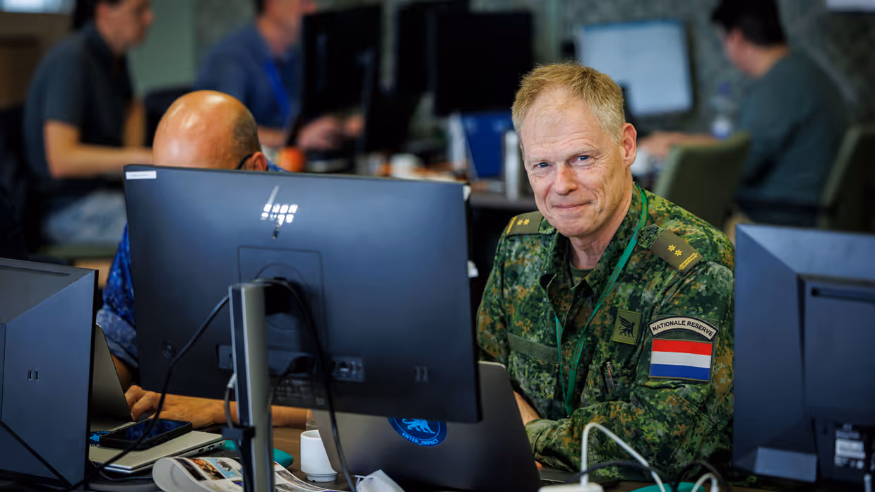 Portretfoto van cyberreservist luitenant-kolonel Arnold Franse, achter een computerscherm met werkende collega’s naast hem en op de achtergrond.