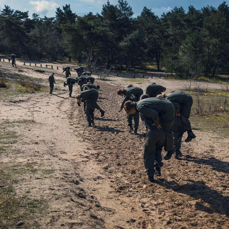 Meer dan 10 militairen rennen over de heide met een collega in de brandweergreep.