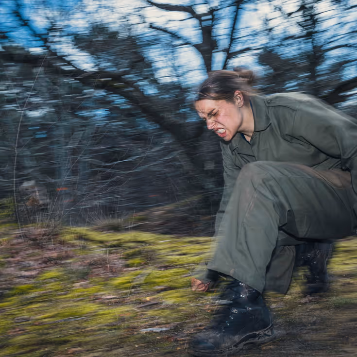 Een vrouwelijke cadet rent met een grimas op gezicht gehurkt door het bos.