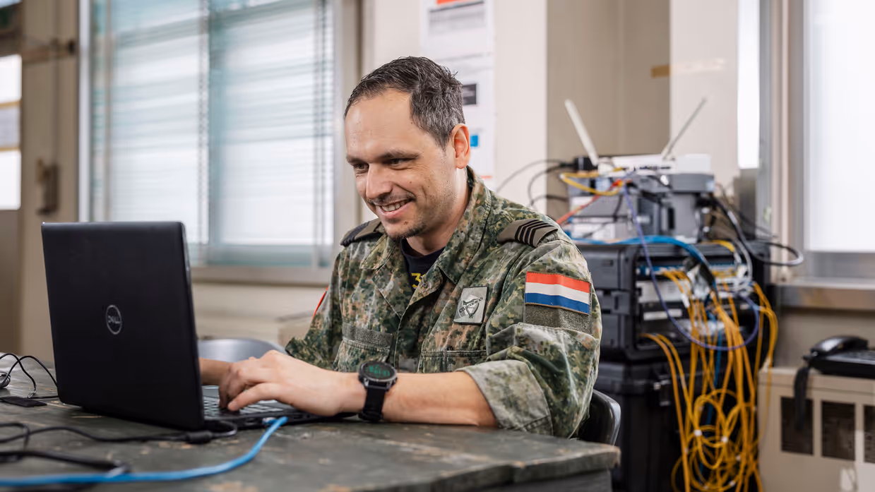: Een militair in camouflagepak zit aan een tafel en werkt op een laptop. In de achtergrond is een wirwar van kabels te zien die uit diverse routers komen.