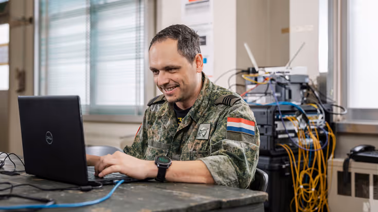 : Een militair in camouflagepak zit aan een tafel en werkt op een laptop. In de achtergrond is een wirwar van kabels te zien die uit diverse routers komen.