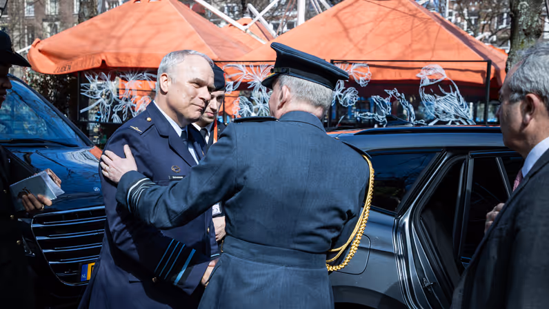  De twee generaals kijken elkaar in de ogen en schudden de hand. Daarachter een zwarte geblindeerde auto.