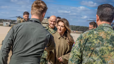 Buiten op een platform schudt de minister de hand van een militair in groene overall. Achter haar staat luitenant-generaal André Steur in gevechtstenue. Om hen heen staan meerdere militairen in gevechtstenue. Op de achtergrond staan Chinooks.