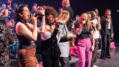 Diverse zangers en zangeressen voor op het podium met in de achtergrond het orkest met dirigent.