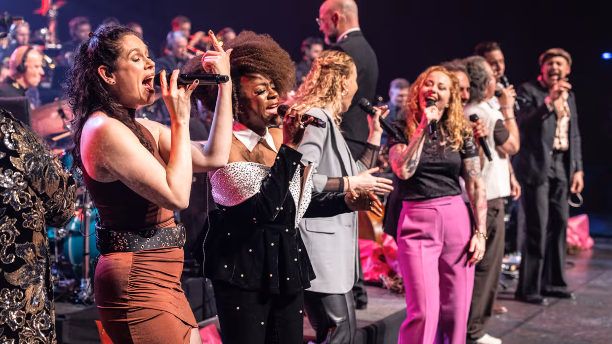 Diverse zangers en zangeressen voor op het podium met in de achtergrond het orkest met dirigent.