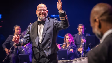 Dirigent majoor Jasper Staps leidt het Orkest Koninklijke Luchtmacht tijdens een concert, terwijl musici op de achtergrond spelen onder podiumverlichting.