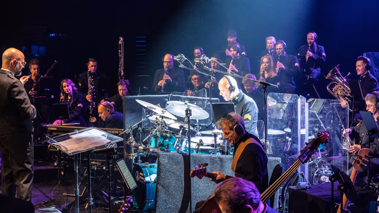 Muzikanten van het Orkest Koninklijke Luchtmacht spelen tijdens een concert, met in het midden fel verlicht de drummer.