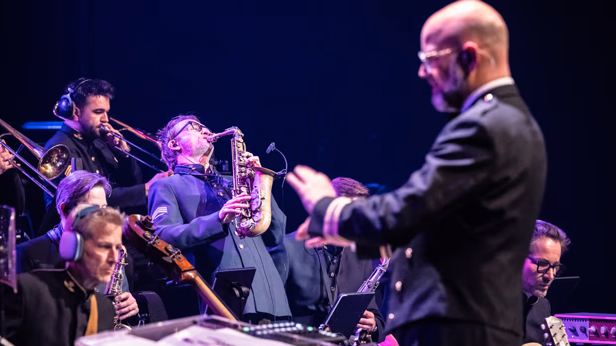 Muzikanten van het Orkest Koninklijke Luchtmacht spelen tijdens een concert, met een saxofonist in solo en de dirigent op de voorgrond.