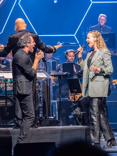 Diverse zangers en zangeressen voor op het podium met in de achtergrond het orkest met dirigent.