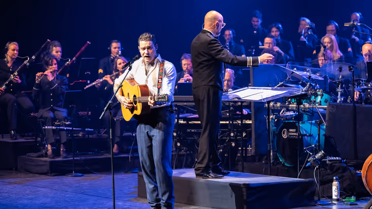 Douwe Bob zingt en speelt gitaar tijdens een concert met het Orkest Koninklijke Luchtmacht, terwijl de dirigent het orkest leidt op de achtergrond.
