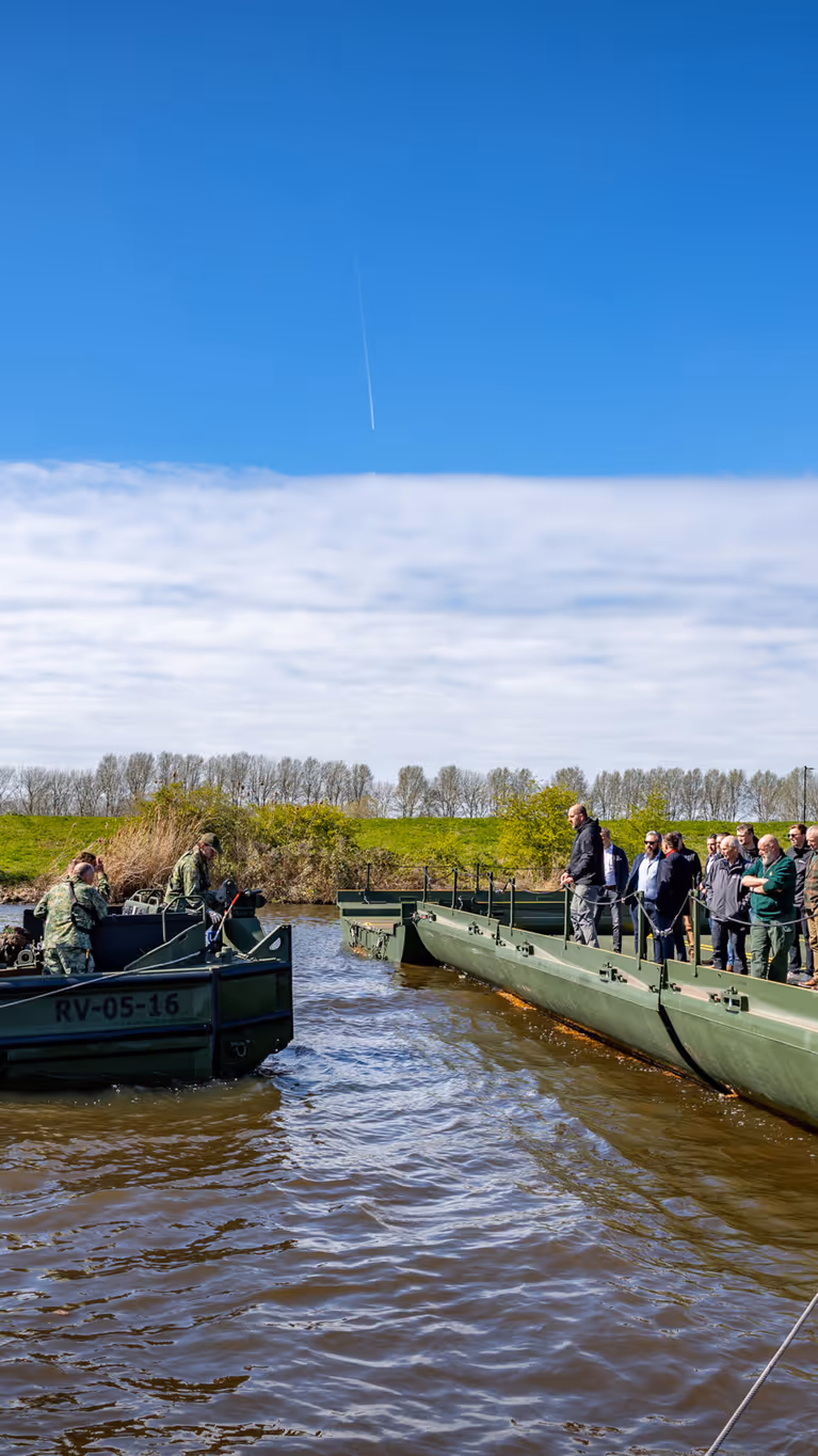 Nieuwe brugslagcapaciteit ligt over een rivier. Daarnaast vaart een boot met militairen. 
