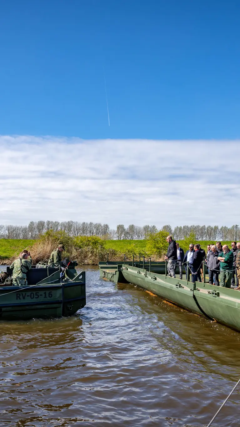 Nieuwe brugslagcapaciteit ligt over een rivier. Daarnaast vaart een boot met militairen. 