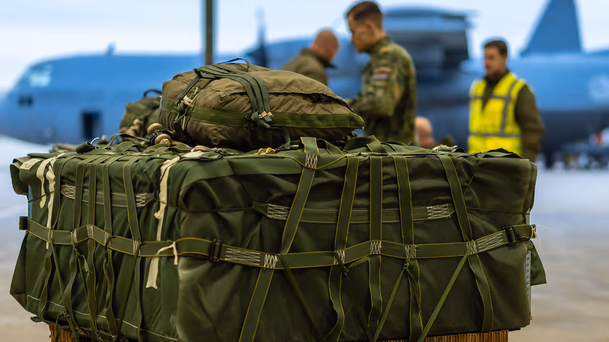 een groene vrachtbundel staat klaar, terwijl op de achtergrond militairen bezig zijn met een ander exemplaar. 