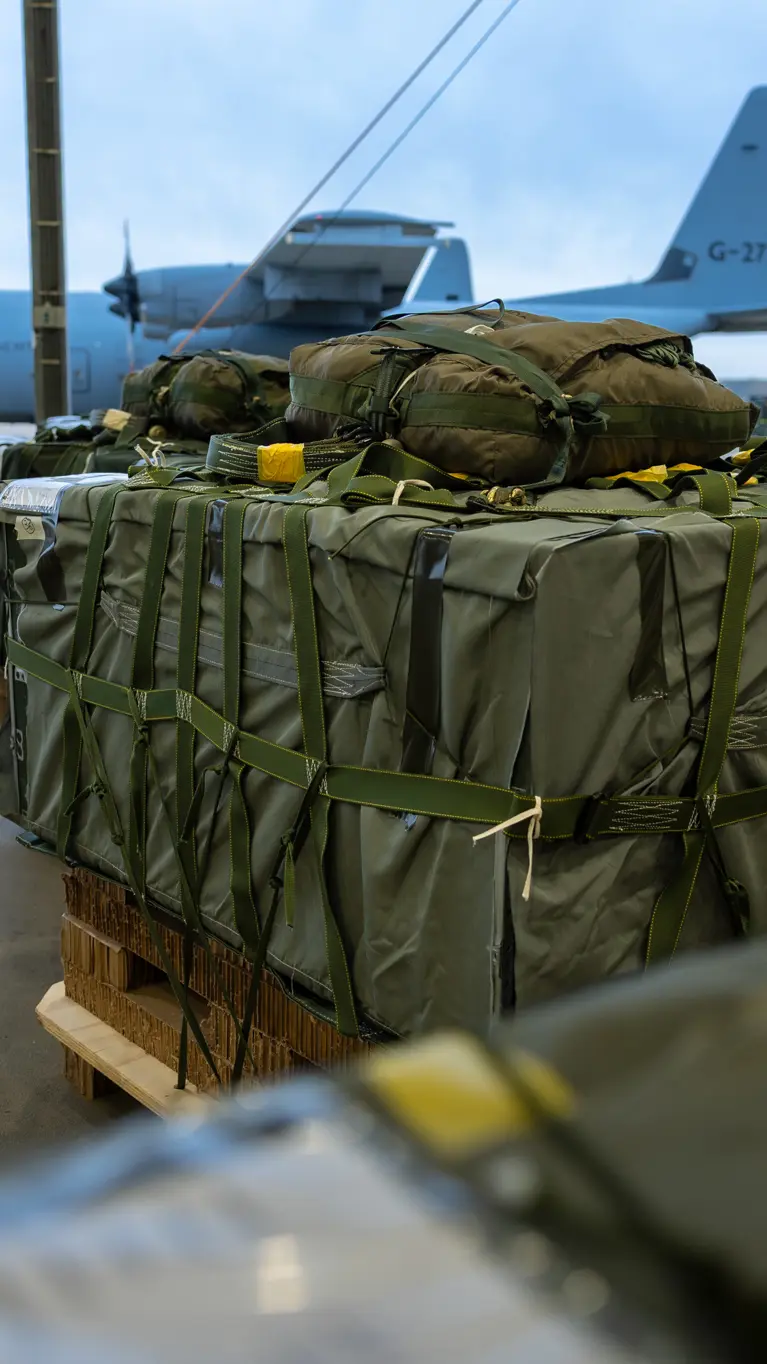 Groene vrachtbundels op pallets. Op de achtergrond staat een grijs transportvliegtuig van de luchtmacht op het platform.