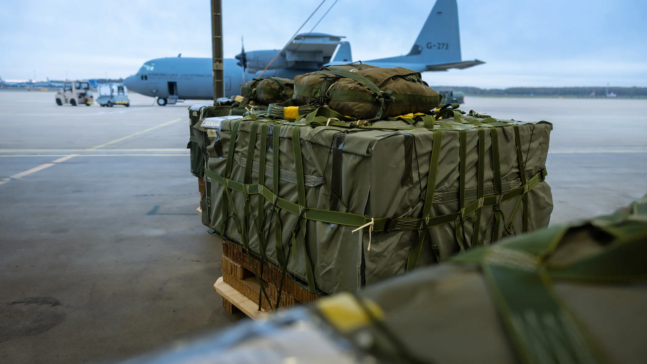 Groene vrachtbundels op pallets. Op de achtergrond staat een grijs transportvliegtuig van de luchtmacht op het platform.