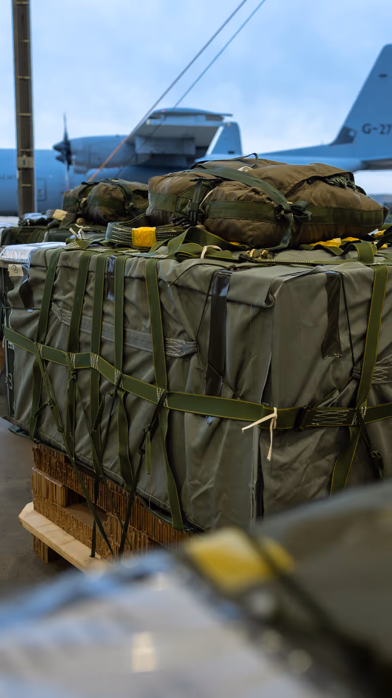 Groene vrachtbundels op pallets. Op de achtergrond staat een grijs transportvliegtuig van de luchtmacht op het platform.