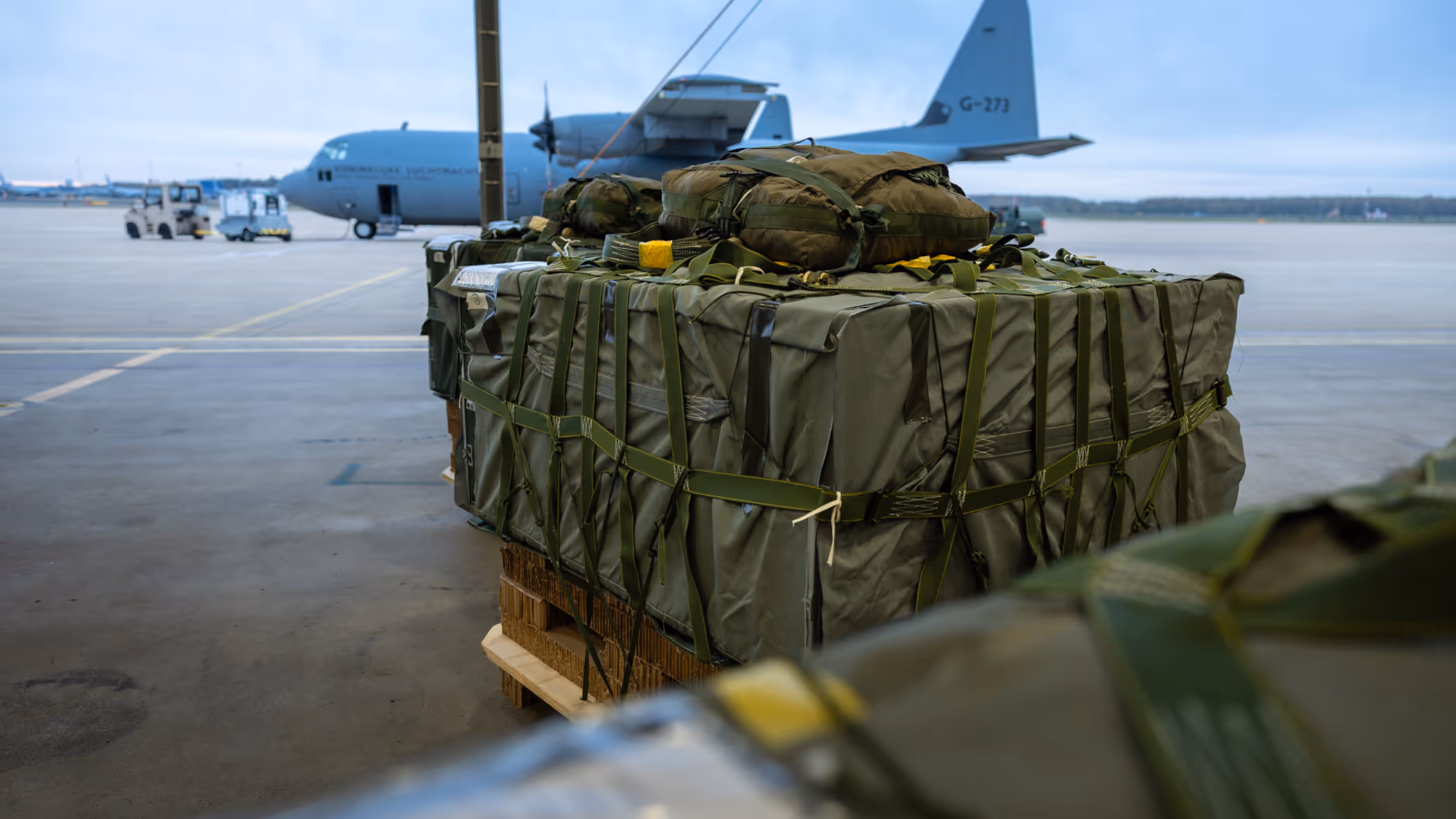 Groene vrachtbundels op pallets. Op de achtergrond staat een grijs transportvliegtuig van de luchtmacht op het platform.