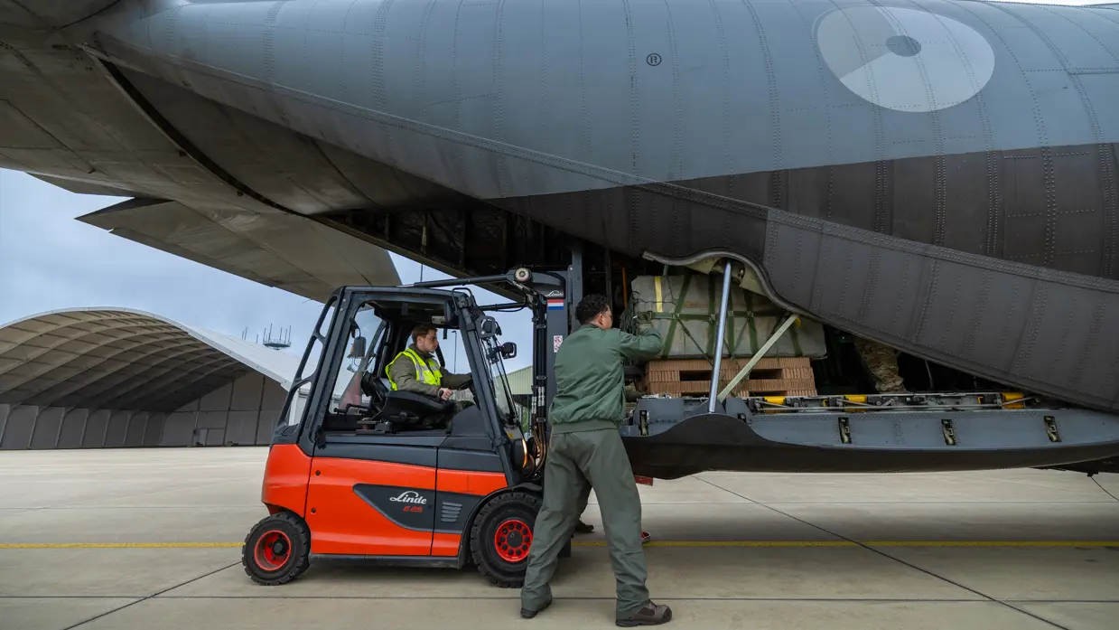 Met een vorkheftruck schuiven 2 militairen vracht in de achterkant van een grijs C-130 transportvliegtuig.