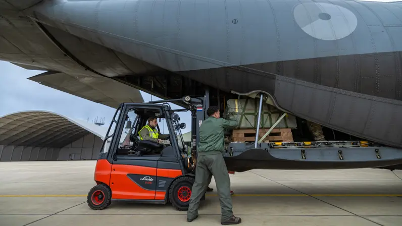 Met een vorkheftruck schuiven 2 militairen vracht in de achterkant van een grijs C-130 transportvliegtuig.